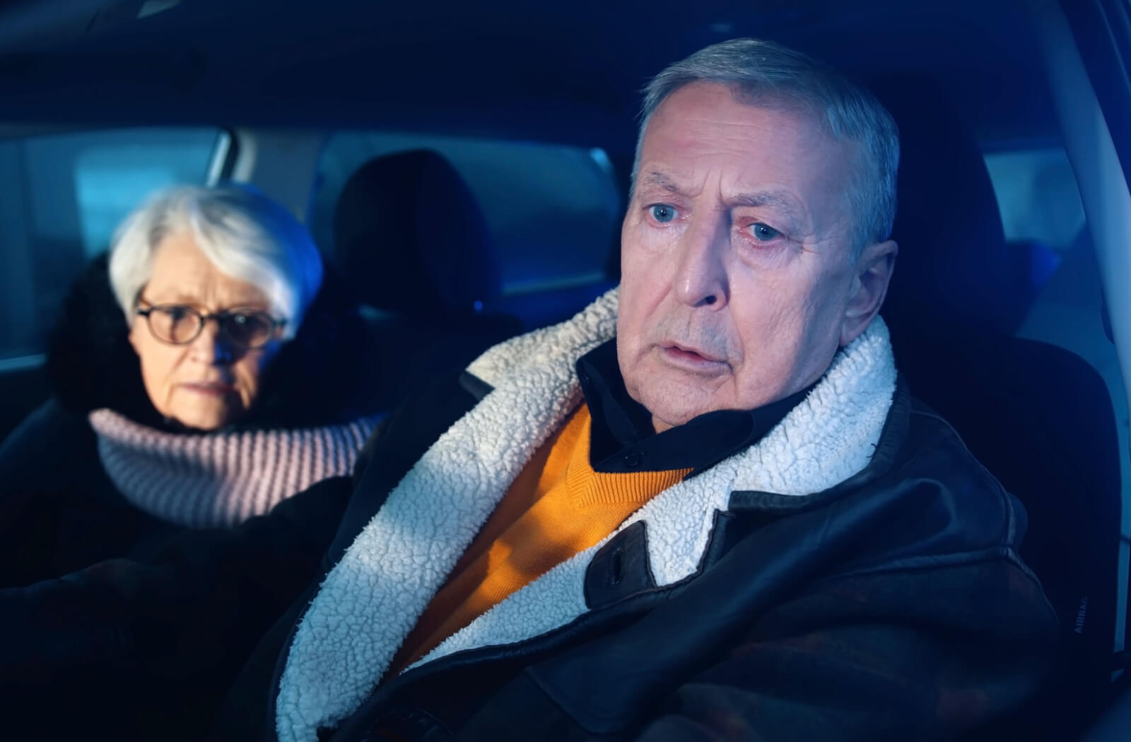 A senior man in the driver's seat of a car with a senior woman in the passenger seat, both with a concerned look on their faces