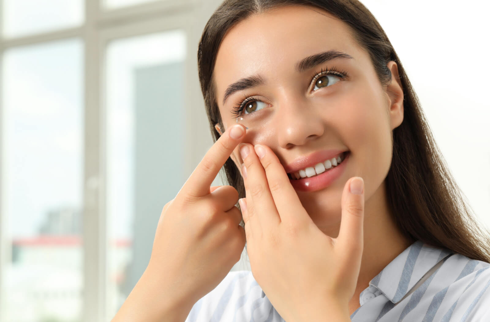 A smiling young woman is inserting a contact lens into her right eye